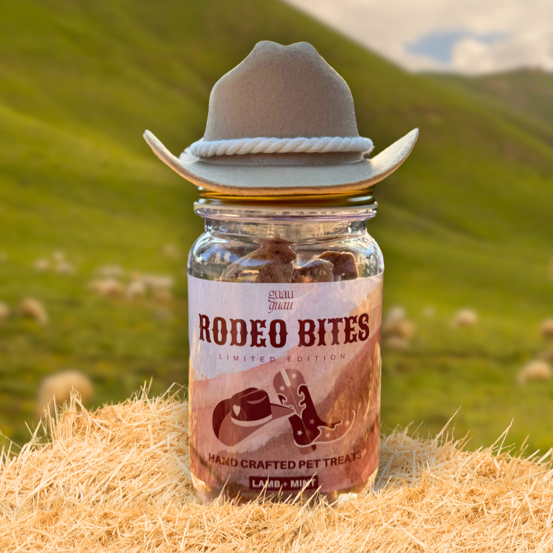 Jar of 'Rodeo Bites' pet treats with a cowboy hat on top, set against a scenic background with green hills and sheep.